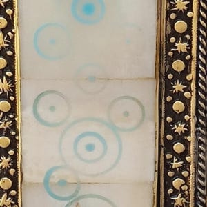 May include: Close-up of a decorative frame with a white center panel featuring light blue concentric circles. The frame has a dark brown border with gold accents, including stars and circular shapes. The design is symmetrical and detailed.