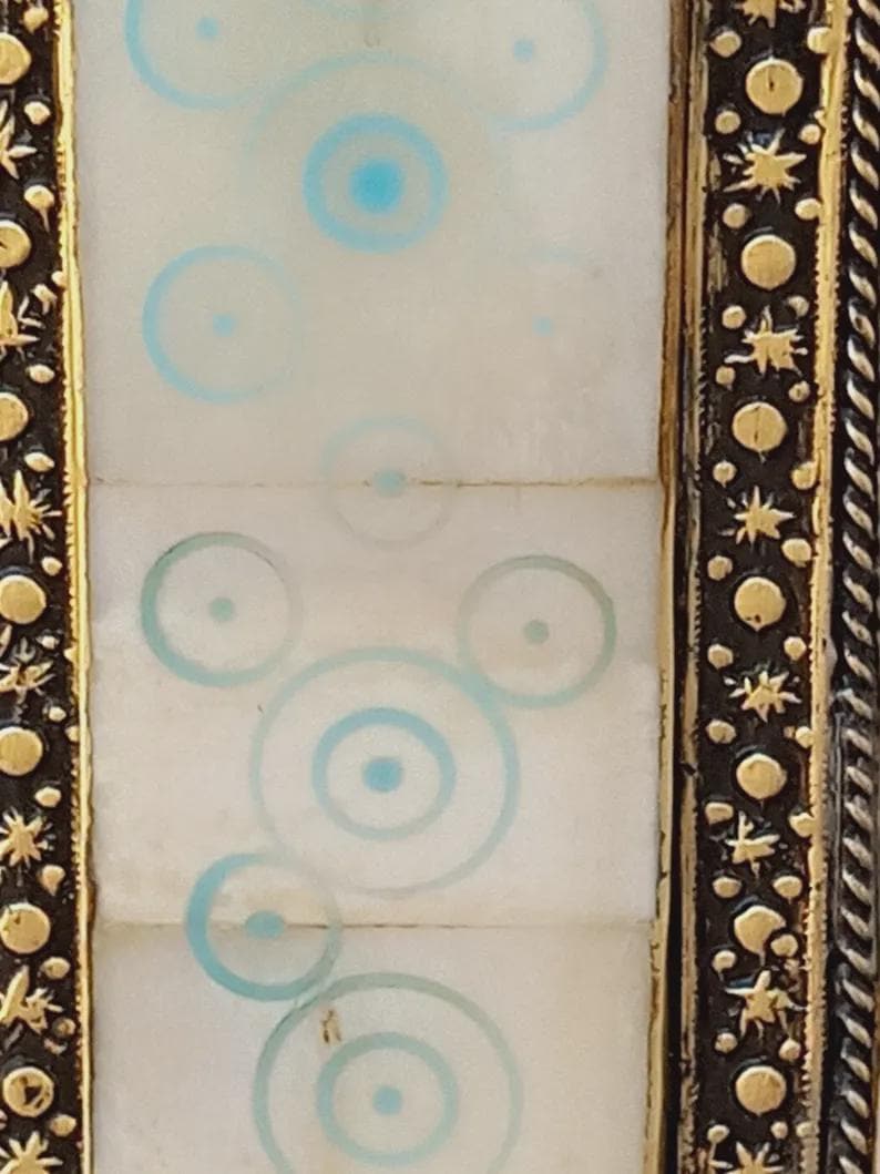 May include: Close-up of a decorative frame with a white center panel featuring light blue concentric circles. The frame has a dark brown border with gold accents, including stars and circular shapes. The design is symmetrical and detailed.