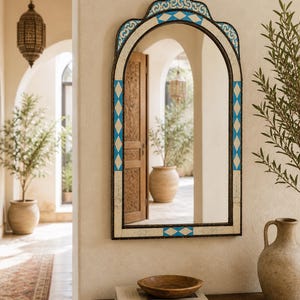 May include: An arched wall mirror with a decorative frame. The frame features a blue and white diamond pattern and a dark border. The mirror is hanging on a cream-colored wall, with a wooden table and decorative items in front of it.