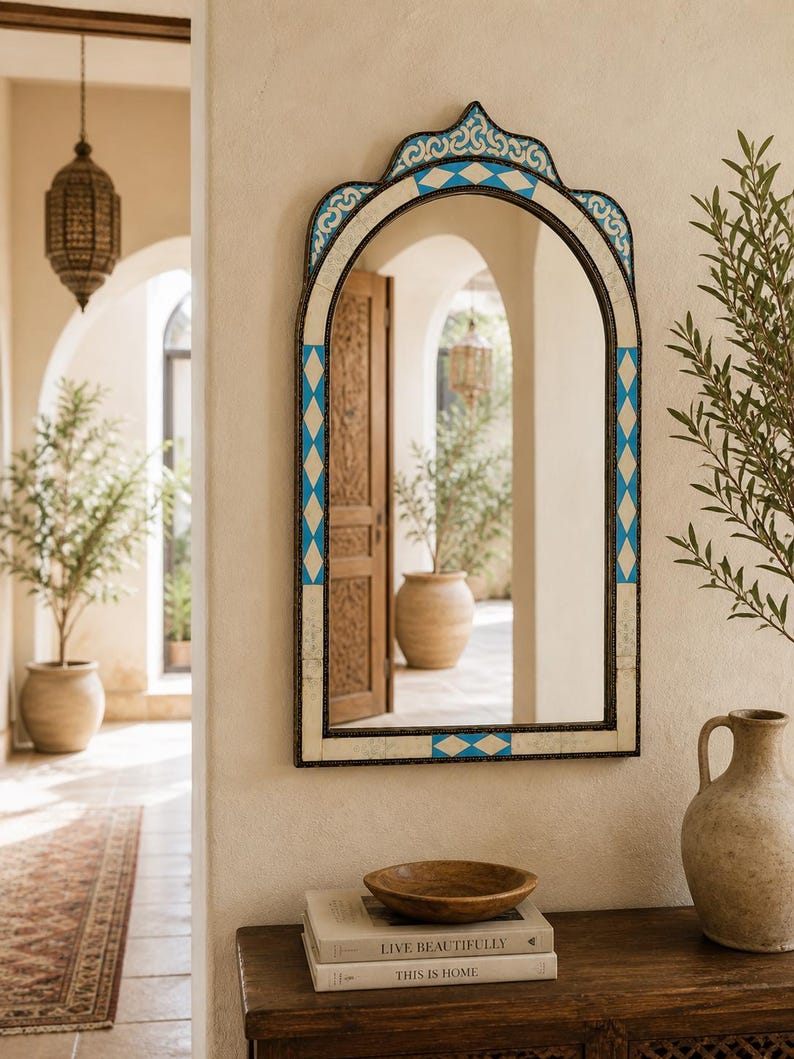 May include: An arched wall mirror with a decorative frame. The frame features a blue and white diamond pattern and a dark border. The mirror is hanging on a cream-colored wall, with a wooden table and decorative items in front of it.