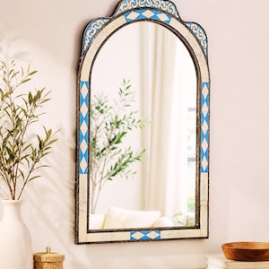 May include: An arched wall mirror with a decorative frame. The frame features a pattern of blue and white diamond shapes and ornate detailing. The mirror is hanging on a white wall, reflecting a room with a white curtain and a wooden table.