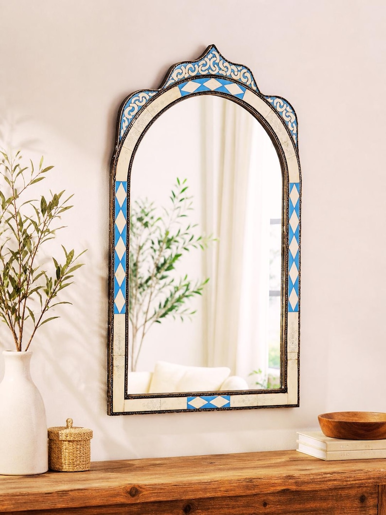 May include: An arched wall mirror with a decorative frame. The frame features a pattern of blue and white diamond shapes and ornate detailing. The mirror is hanging on a white wall, reflecting a room with a white curtain and a wooden table.