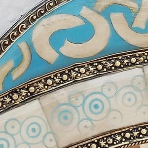 May include: Close-up of a decorative object with a turquoise and white design. The object has a silver-colored metal border with intricate detailing. The turquoise sections feature white curved shapes, while the white sections have blue concentric circles.