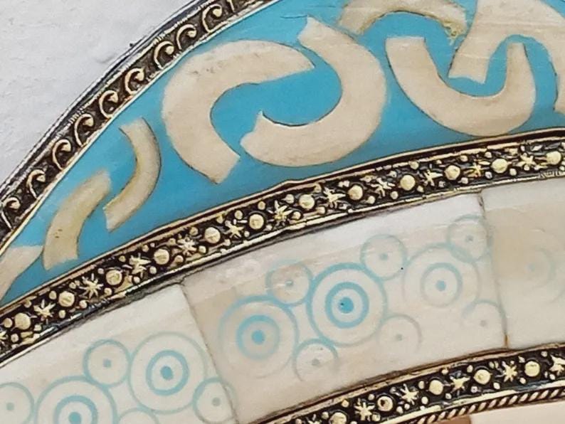 May include: Close-up of a decorative object with a turquoise and white design. The object has a silver-colored metal border with intricate detailing. The turquoise sections feature white curved shapes, while the white sections have blue concentric circles.