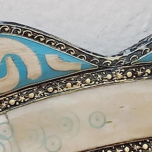 May include: Close-up of a decorative object with a cream-colored surface and a border featuring intricate gold and black detailing. The design includes sections of light blue and white shapes, with a pattern of small blue circles.