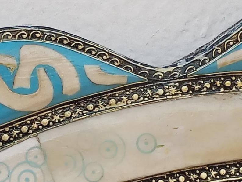 May include: Close-up of a decorative object with a cream-colored surface and a border featuring intricate gold and black detailing. The design includes sections of light blue and white shapes, with a pattern of small blue circles.