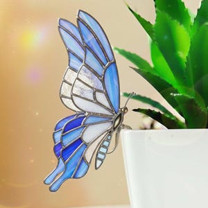 May include: A stained glass butterfly with blue and white wings. The butterfly is perched on the edge of a white pot with a green plant. The wings have a detailed design with various shades of blue.