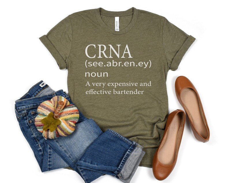 Op de afbeelding: Een groen T-shirt met witte tekst die "CRNA (see.abr.en.ey) noun A very expensive and effective bartender" luidt. Het shirt ligt op een wit oppervlak met een paar bruine ballerina's en een blauwe spijkerbroek met een kleine gebreide pompoen.