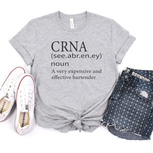 Op de afbeelding: Een grijs T-shirt met de tekst "CRNA (see.abr.en.ey) noun A very expensive and effective bartender" erop gedrukt. Het shirt is onderaan geknoopt en wordt getoond met een paar witte sneakers en een blauwe spijkerbroek met een wit stippenpatroon.