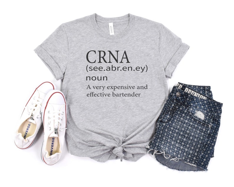 Op de afbeelding: Een grijs T-shirt met de tekst "CRNA (see.abr.en.ey) noun A very expensive and effective bartender" erop gedrukt. Het shirt is onderaan geknoopt en wordt getoond met een paar witte sneakers en een blauwe spijkerbroek met een wit stippenpatroon.