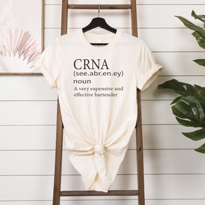 Op de afbeelding: Een beige T-shirt met de tekst "CRNA (see.abr.en.ey) noun A very expensive and effective bartender" gedrukt op de voorkant.