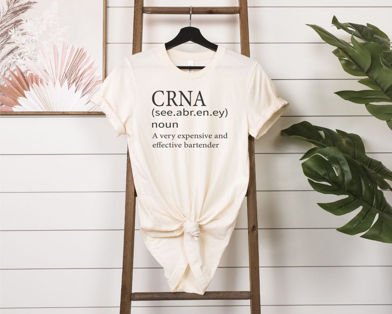 Op de afbeelding: Een beige T-shirt met de tekst "CRNA (see.abr.en.ey) noun A very expensive and effective bartender" gedrukt op de voorkant.