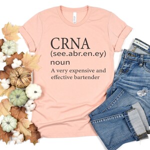 Op de afbeelding: Een roze T-shirt met de tekst "CRNA (see.abr.en.ey) noun A very expensive and effective bartender" erop gedrukt.