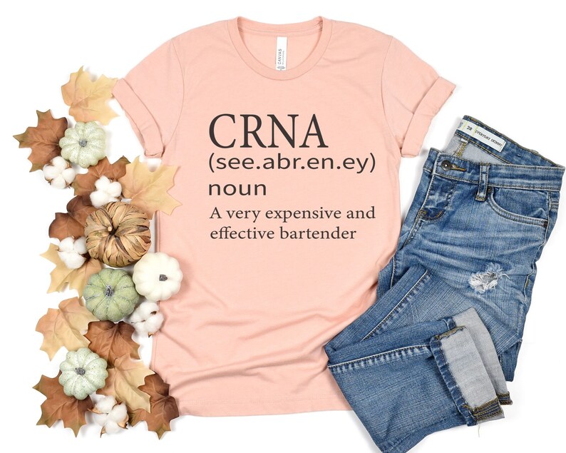 Op de afbeelding: Een roze T-shirt met de tekst "CRNA (see.abr.en.ey) noun A very expensive and effective bartender" erop gedrukt.