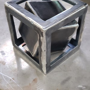 K&ouml;nnte beinhalten: Eine Metall-Kubussculptur mit einem kleineren W&uuml;rfel im Inneren. Die Skulptur ist aus geschwei&szlig;tem Stahl gefertigt und hat einen rohen, industriellen Look.