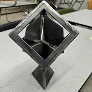 K&ouml;nnte beinhalten: Eine Metallskulptur eines W&uuml;rfels in einem Diamanten. Die Skulptur ist aus dunklem Metall gefertigt und hat eine gl&auml;nzende Oberfl&auml;che. Sie steht auf einer grauen Oberfl&auml;che.