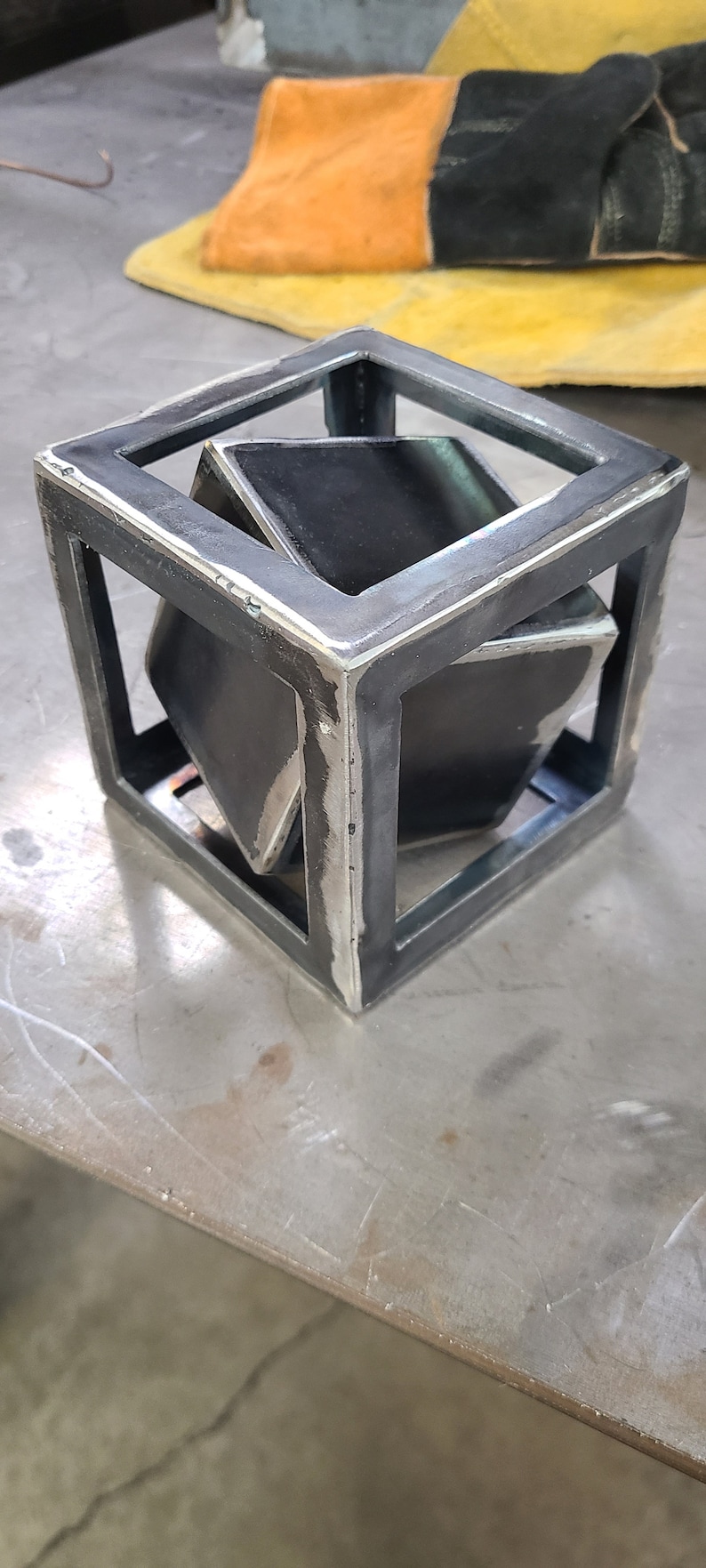 K&ouml;nnte beinhalten: Eine Metall-Kubussculptur mit einem kleineren W&uuml;rfel im Inneren. Die Skulptur ist aus geschwei&szlig;tem Stahl gefertigt und hat einen rohen, industriellen Look.