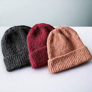 May include: Three knitted beanies in different colors: gray, burgundy, and light pink. The beanies are folded and arranged on a white surface.