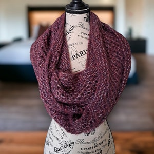 May include: A maroon infinity scarf with a textured knit pattern. The scarf is displayed on a mannequin with a vintage Parisian design.