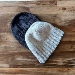 Peut inclure: Deux bonnets en tricot, l'un noir et l'autre beige, sont empilés l'un sur l'autre. Les bonnets sont faits d'un fil doux et texturé.