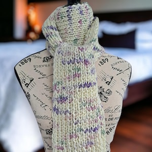 Peut inclure: Écharpe tricotée de couleur crème avec des mouchetures violettes, vertes et roses. L'écharpe est présentée sur un mannequin. L'écharpe en tricot épais est un accessoire douillet pour le temps froid. L'écharpe est faite à la main.