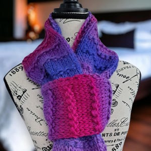 May include: A hand-knitted scarf in shades of purple, blue, and pink, displayed on a mannequin. The scarf features a textured cable knit design and is made of soft yarn. The scarf is a cozy accessory for cold weather.