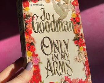 Op de afbeelding: Een vintage paperback boek getiteld "Only In My Arms" van Jo Goodman. De omslag heeft een bloemenrand met rode, roze en gele bloemen en twee witte duiven. De titel en de naam van de auteur zijn in gouden letters.