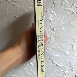 May include: A paperback copy of "The Notebook" by Nicholas Sparks. The book's spine is cream-colored with blue text and the Warner Vision logo. The book is a New York Times bestseller and is labeled as fiction.
