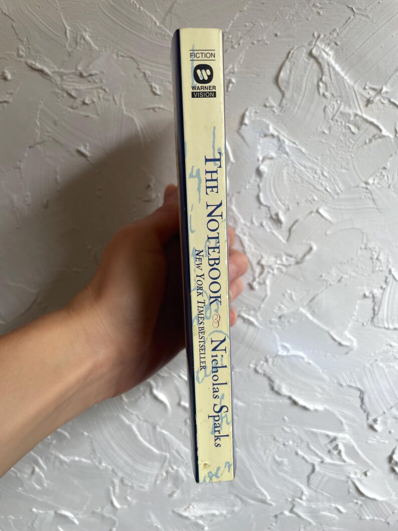 May include: A paperback copy of "The Notebook" by Nicholas Sparks. The book's spine is cream-colored with blue text and the Warner Vision logo. The book is a New York Times bestseller and is labeled as fiction.
