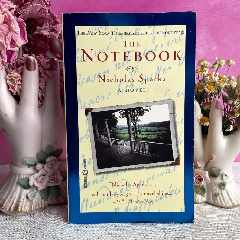 May include: A paperback copy of "The Notebook" by Nicholas Sparks. The book has a blue border and a photo of a porch on the cover. The title is in red, and the author's name is in black.