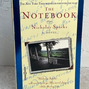 May include: A hardcover copy of "The Notebook" by Nicholas Sparks. The book has a blue border and a cream-colored cover with the title in large red letters. A small photo of a porch with a scenic view is featured.