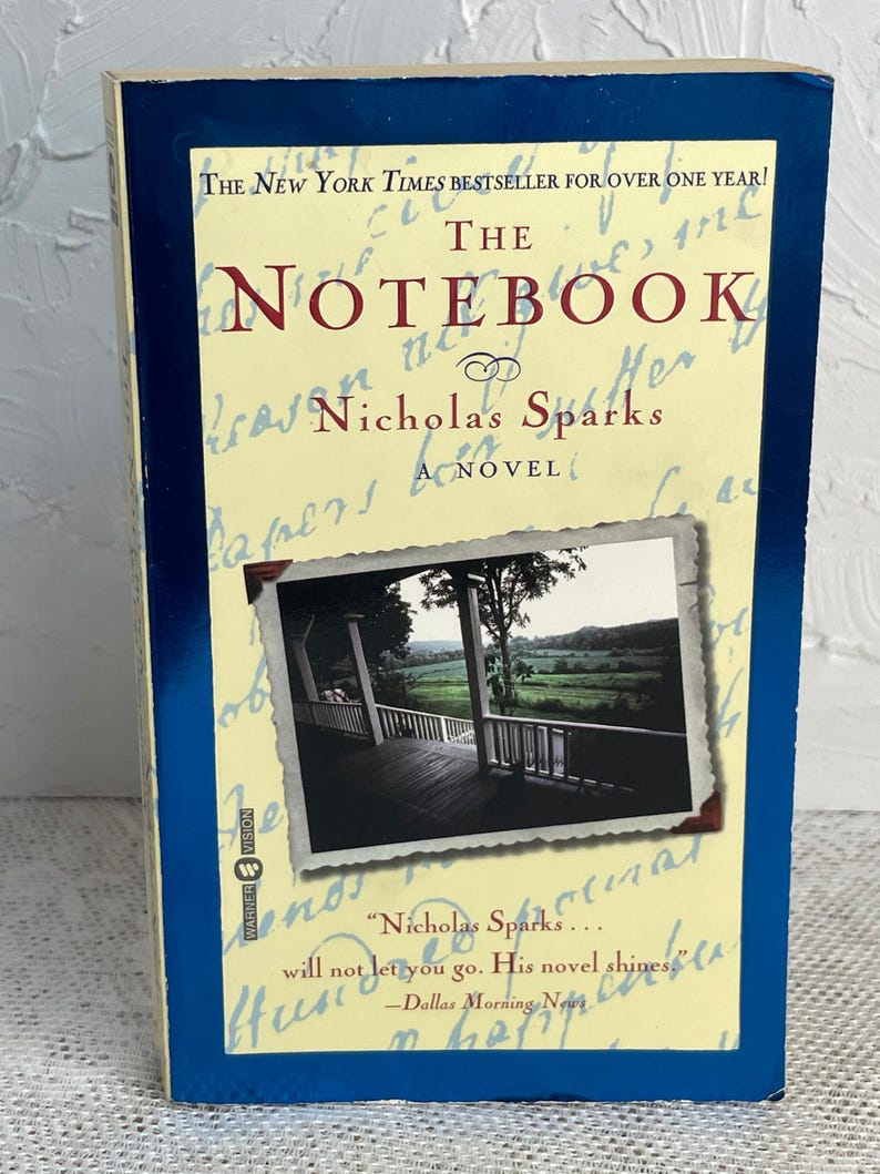May include: A hardcover copy of "The Notebook" by Nicholas Sparks. The book has a blue border and a cream-colored cover with the title in large red letters. A small photo of a porch with a scenic view is featured.