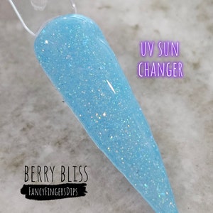 May include: A light blue nail swatch with glitter, labeled "UV Sun Changer." The swatch is from "Berry Bliss" by "Fancy Fingers Dips." The nail is a long, pointed shape, showcasing the color and glitter effect.