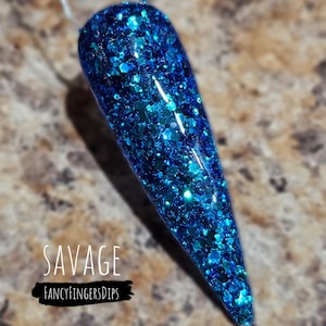 May include: A blue glitter nail swatch with the word "SAVAGE" in white text and the brand name "FANCYFINGERSDIPS" in a black banner. The nail is a long, pointed shape, covered in sparkling blue glitter.