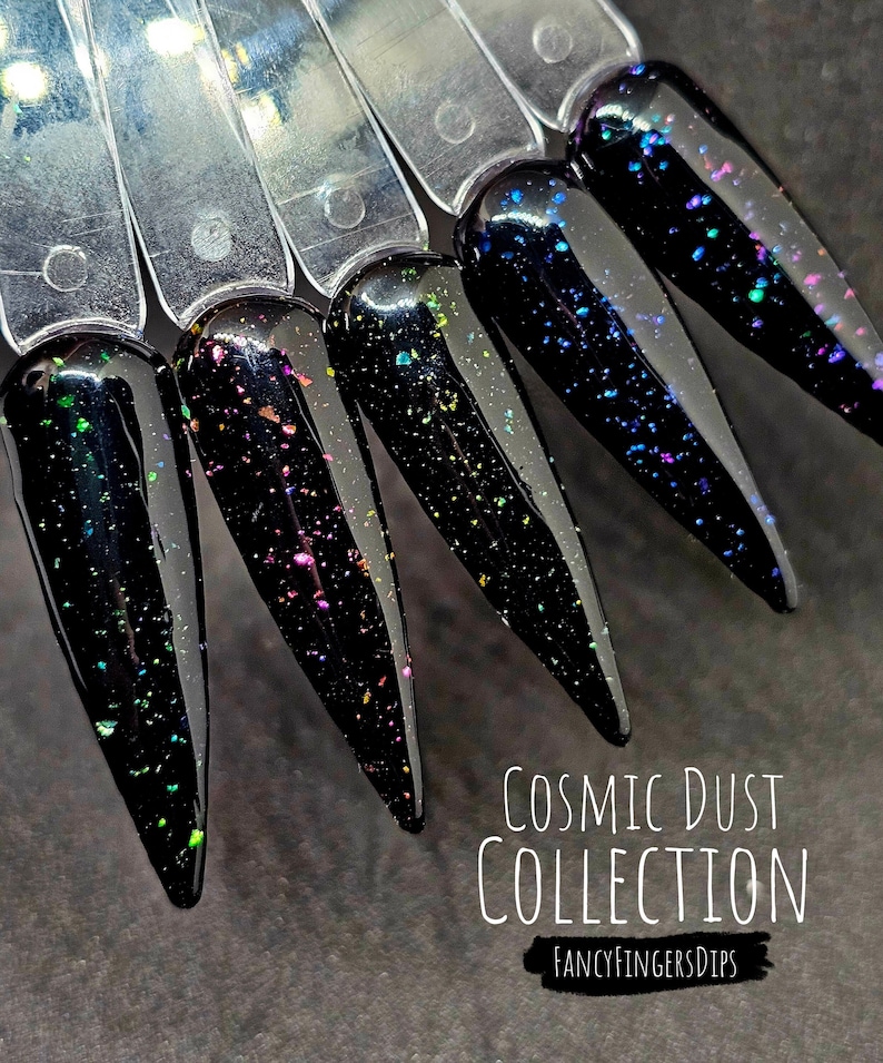 May include: Five black nail dip powder colours with iridescent glitter in a variety of colours. The colours are displayed on a clear plastic fan. The text "COSMIC DUST COLLECTION" and "FANCYFINGERSDIPS" is displayed below the nail dips.