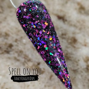 May include: A close-up of a black and purple glitter nail dip powder with iridescent sparkles. The text "SPELL ON YOU" and "FANCYFINGERSDIPS" is visible on the image.
