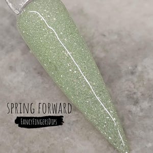 May include: A light green nail polish swatch with glitter, labeled "Spring Forward" and "Fancy Fingers Dips". The polish is applied to a clear plastic stick, showcasing the color and texture. The background is a light gray surface.