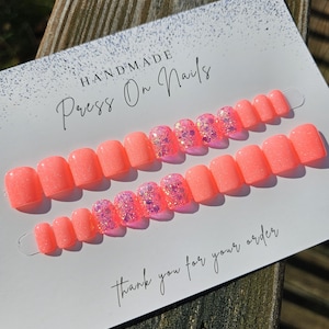 Könnte beinhalten: Zwei Sets handgefertigter Press-On-Nägel in leuchtendem Korallenrot. Einige Nägel weisen ein Glitzerdesign mit rosa und lila Glitzer auf. Auf der Karte steht "HANDMADE Press On Nails" und "Thank you for your order".