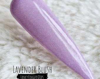 Puede incluir: Una muestra de esmalte de uñas de color lavanda con un acabado brillante. El texto "Lavender Blush" se muestra encima de una pancarta negra que dice "Fancy Fingers Dips". La muestra está sobre una superficie blanca y esponjosa.