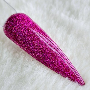 May include: A vibrant, magenta-colored nail with a glittery finish. The nail is long and pointed, with a smooth, glossy surface. The glitter particles catch the light, creating a sparkling effect. The nail is displayed against a soft, white background.