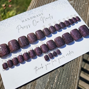 Peut inclure: Un ensemble de faux ongles faits à la main, de couleur prune foncée avec une finition pailletée. Les ongles sont disposés sur une carte blanche avec le texte "HANDMADE Press On Nails" et "Thank you for your order."