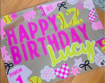 May include: A rectangular poster on brown paper with the text HAPPY 12TH BIRTHDAY Lucy. The design features bright pink and neon yellow lettering, softball graphics, a jersey with the name FOUCHE and number 1, hearts, stars, and bows.