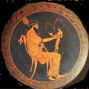 May include: Ancient Greek pottery plate with a black background and orange figures. The central image depicts a seated figure holding a cup, with another figure standing before him. The plate has a decorative border with a repeating geometric pattern.