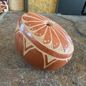 May include: A reddish-brown, oval-shaped decorative object with carved floral and geometric patterns. The object has a hole at the top and is likely made of ceramic or a similar material. The carved designs are a lighter color than the base.