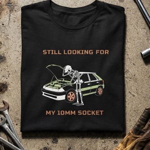 Puede incluir: Una camiseta negra con el gráfico de un esqueleto mirando bajo el capó de un coche y el texto "STILL LOOKING FOR MY 10MM SOCKET" en naranja. El diseño incluye un coche negro con detalles en verde y ruedas de color naranja.