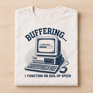May include: A cream-colored t-shirt featuring a dark blue graphic of a vintage computer with a loading bar on the screen. The text reads "BUFFERING..." above the computer and "I FUNCTION ON DIAL-UP SPEED" below the keyboard and mouse illustration.