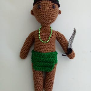 May include: A crocheted doll with brown skin, wearing a green and black hat, a green skirt, and a green beaded necklace. The doll is holding a silver knife in its right hand.