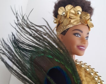 May include: A black doll with brown hair wearing a gold crown with gold flowers and a gold necklace. The doll is holding a peacock feather fan.