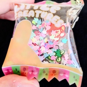 May include: A clear plastic bag filled with colorful confetti and a cartoon character. The bag has the words "LITTLE CREATURES" printed on it. The confetti includes shapes of bunnies, flowers, and other small figures. The bottom of the bag has a colorful striped design.