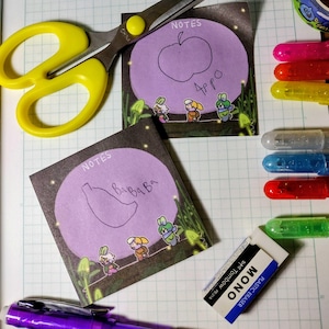 Può includere: Due post-it quadrati con disegni di una banana e una mela, ciascuno all'interno di un cerchio viola, con la parola "NOTES" sopra. Un paio di forbici gialle, penne colorate e una gomma sono anche nell'immagine.